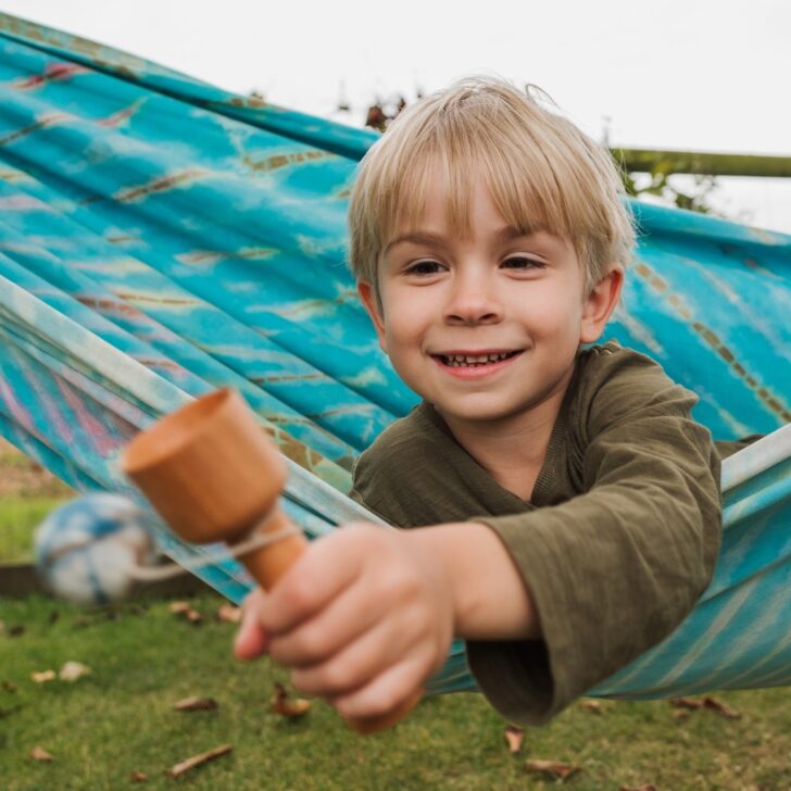 A young boy plays with a wooden toy while relaxing in a hammock, surrounded by greenery