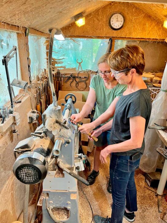 Liz and a woman collaboratively operating a woodworking machine in a workshop setting
