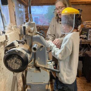 Girl uses a woodturning lathe under the guidance of a teacher.