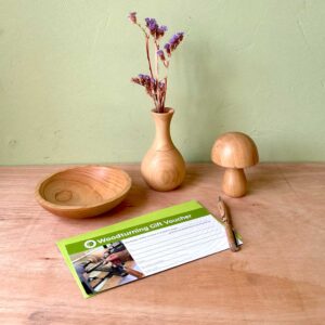 Love Heartwood woodturning gift voucher with green envelope lying next to a handmade wooden bowl, pen, vase and mushroom.