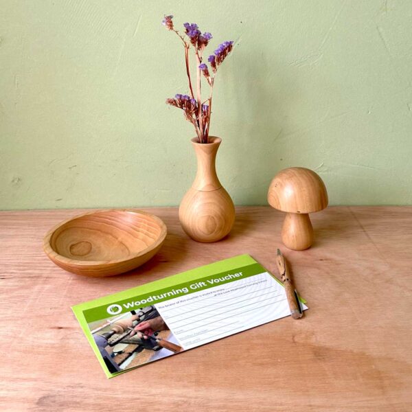 Love Heartwood woodturning gift voucher with green envelope lying next to a handmade wooden bowl, pen, vase and mushroom.