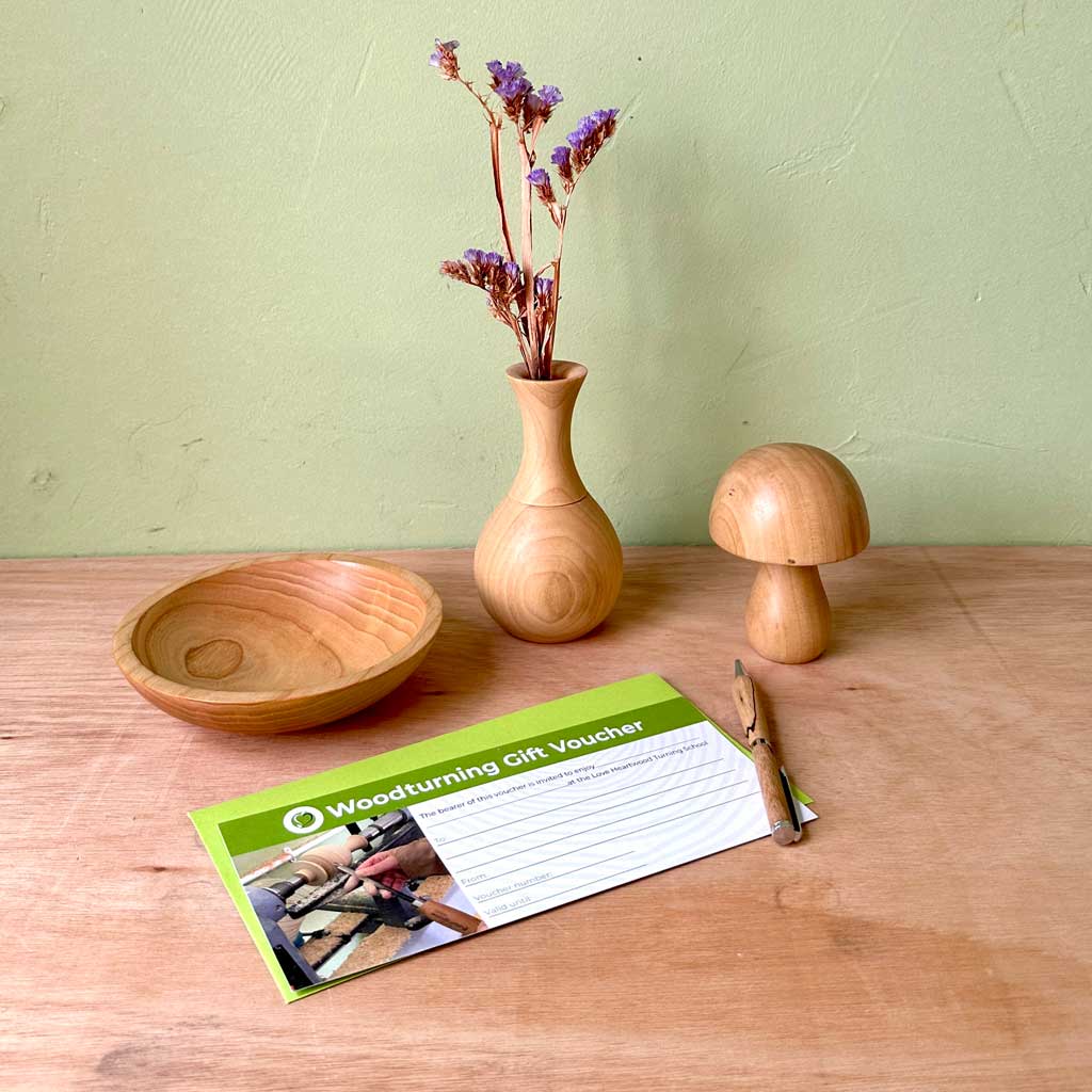 Love Heartwood woodturning gift voucher with green envelope lying next to a handmade wooden bowl, pen, vase and mushroom.