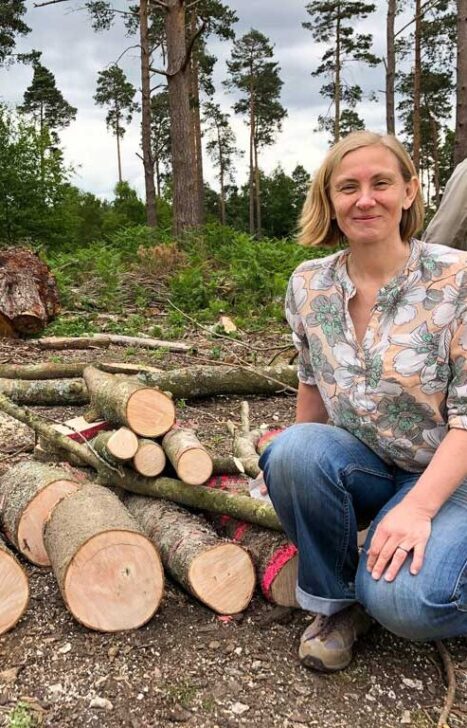 Liz kneeling beside stacked logs in a serene forest setting, surrounded by trees and natural foliage