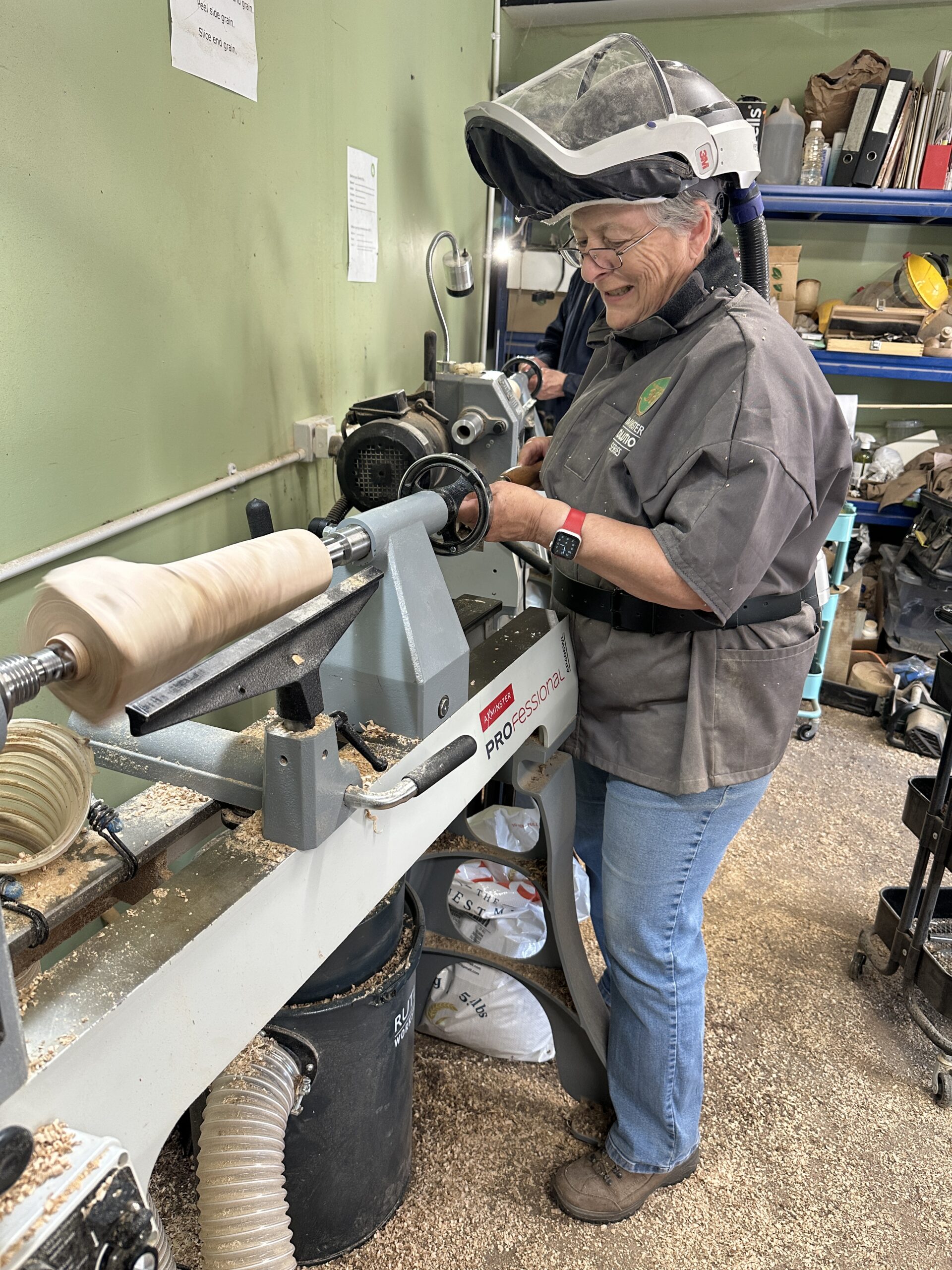 Turner standing at the end of a woodturning lathe and drilling into the end of a wooden table lamp.