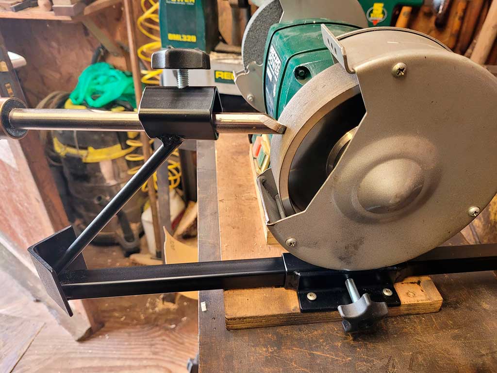 A black metal base slide and jig holding a bowl gouge against the wheel of a bench grinder to sharpen it.