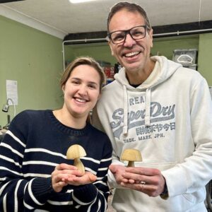 A smiling father and daughter hold their unique wooden mushrooms up to the camera.